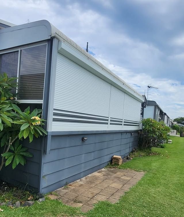 A blue and gray mobile home with a white shutter, on a green lawn under a cloudy sky.