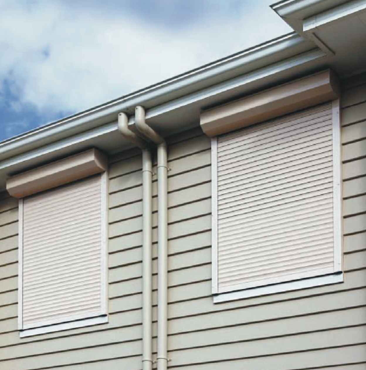 Two beige roll-down shutters covering windows on a beige siding house, under a gutter.