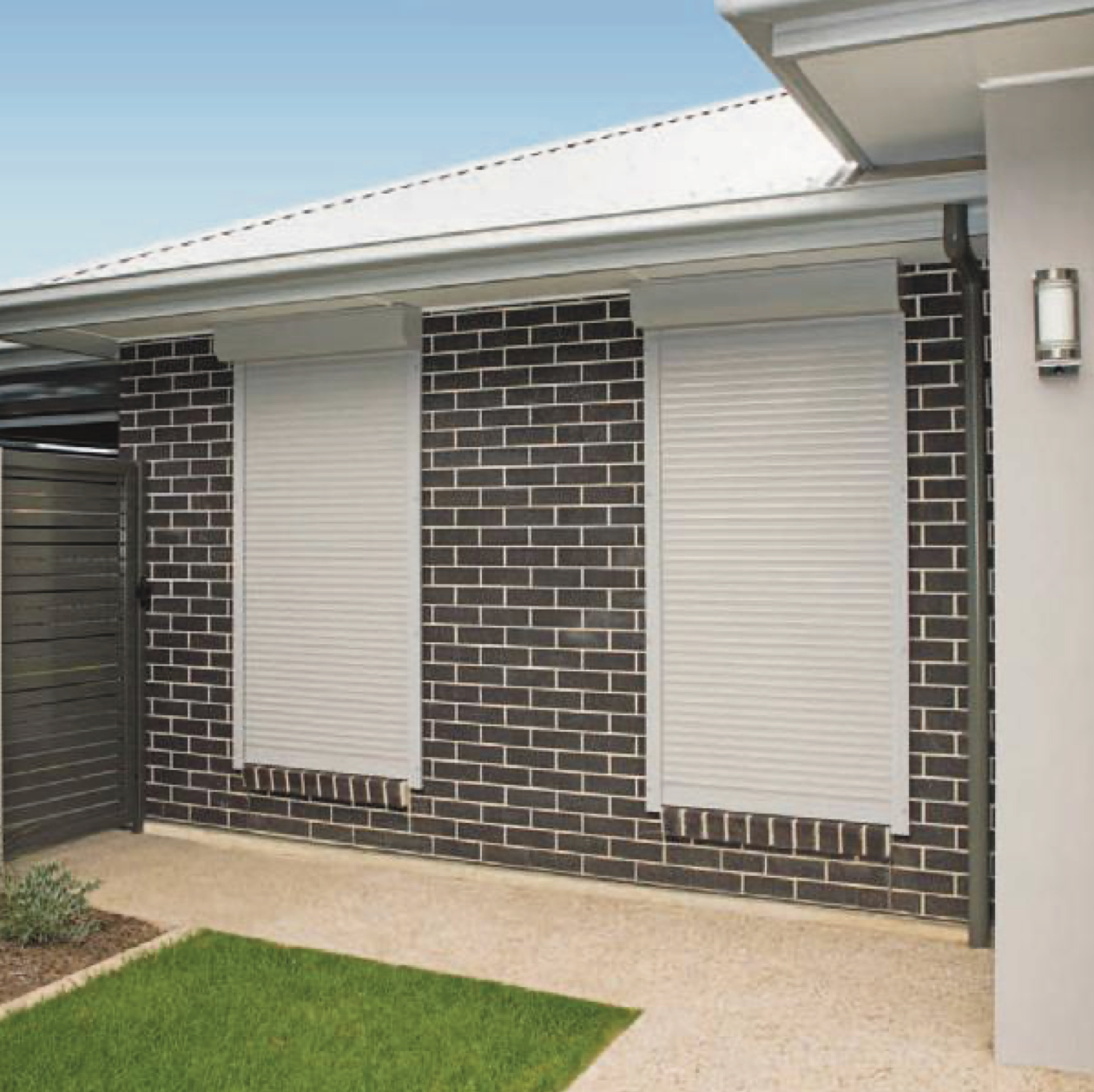 Two white roller shutters on dark brick wall of house, with a green patch of grass.