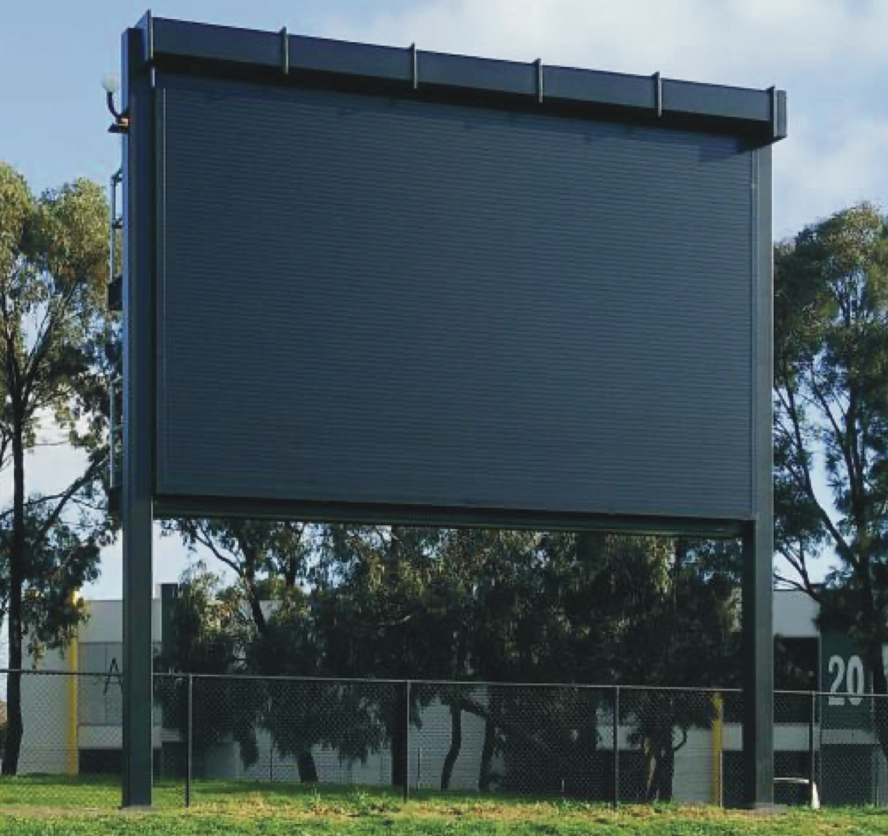 Large, black scoreboard on metal posts in a grassy field, trees in the background, a building with a