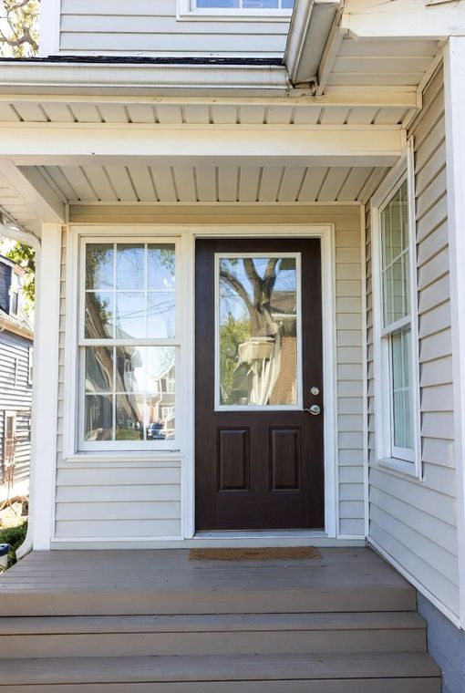 Brown front door with glass panel flanked by two windows on a light-colored house porch.