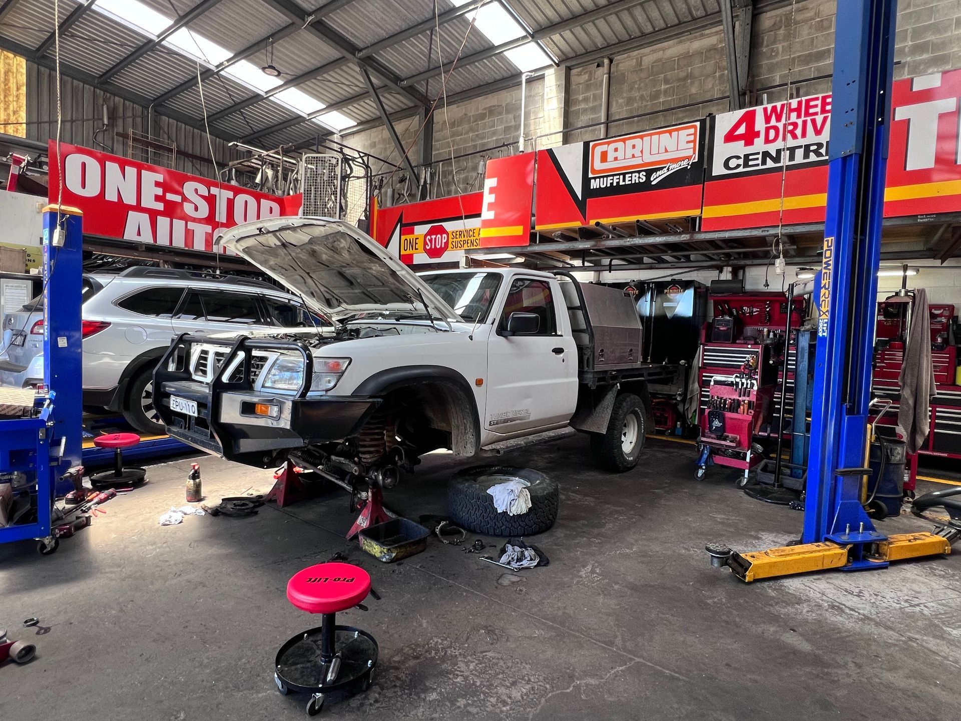 A mechanic shop with a white pickup truck on a lift. Tools and parts are scattered around. — One Stop Service and Suspension Centre In Port Macquarie, NSW