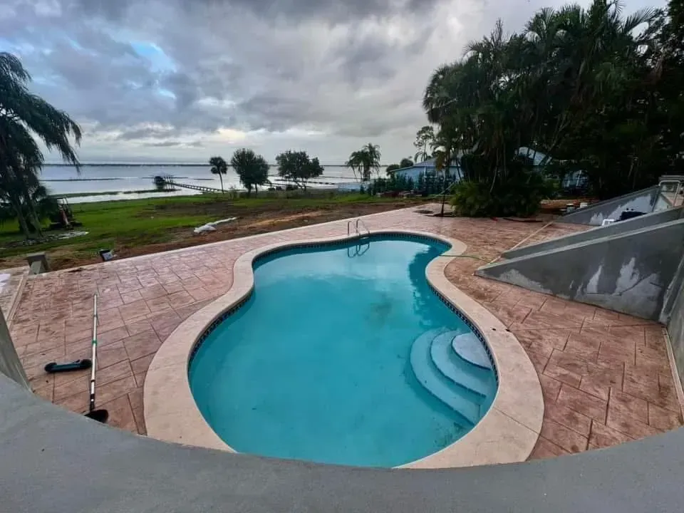 A large swimming pool is surrounded by palm trees and a view of the ocean.