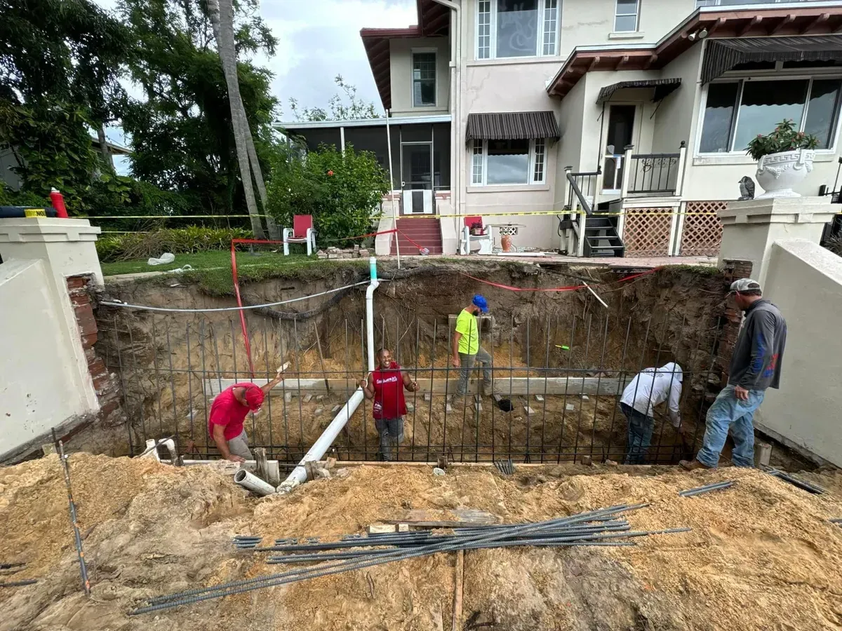 A group of construction workers are working on a large hole in the ground in front of a house.