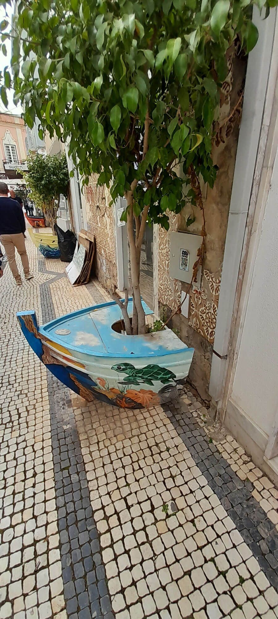 Auf dem Bürgersteig neben einem Baum liegt ein bemaltes Boot.