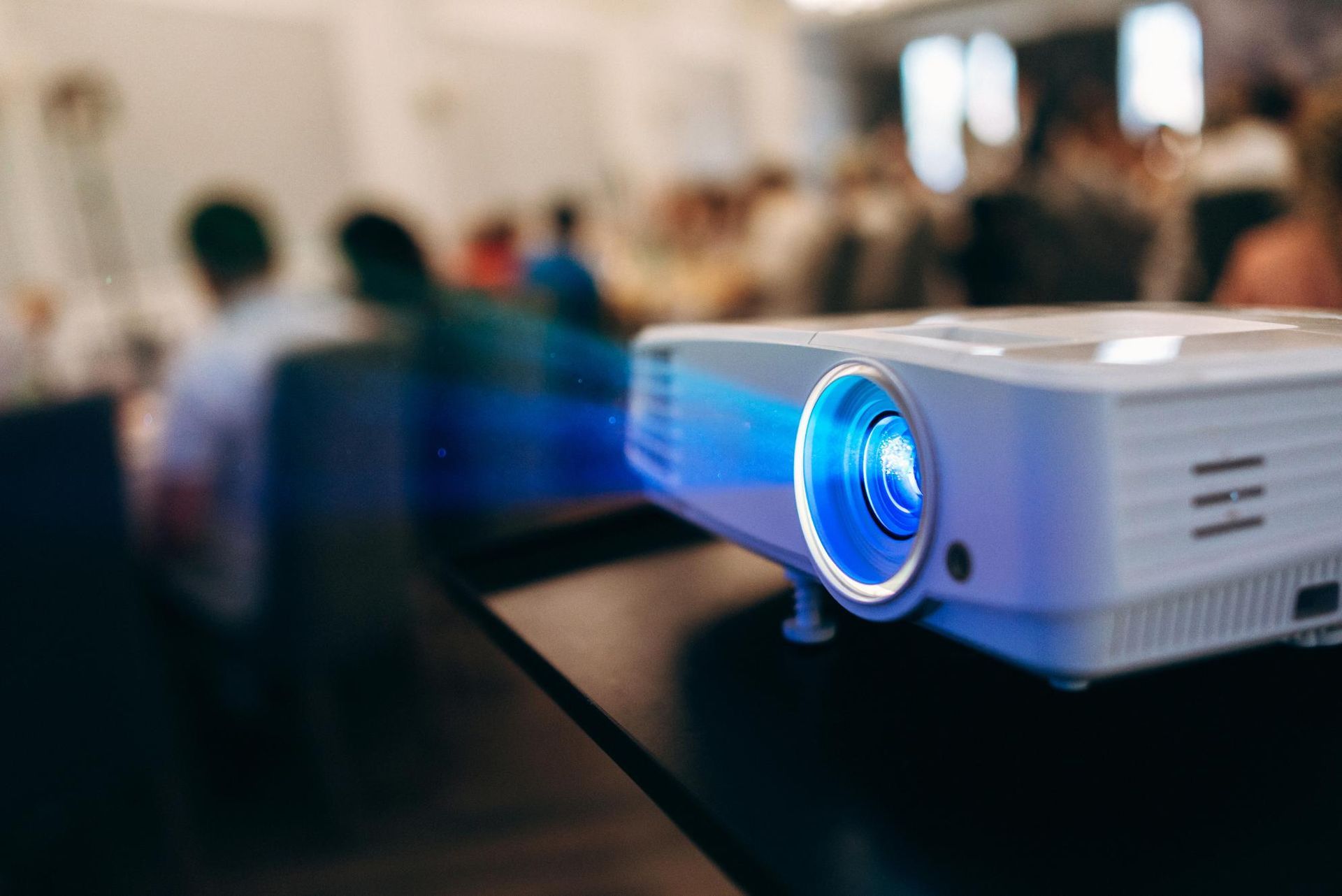 Blue-lit projector on a table in a blurred conference room with people in the background