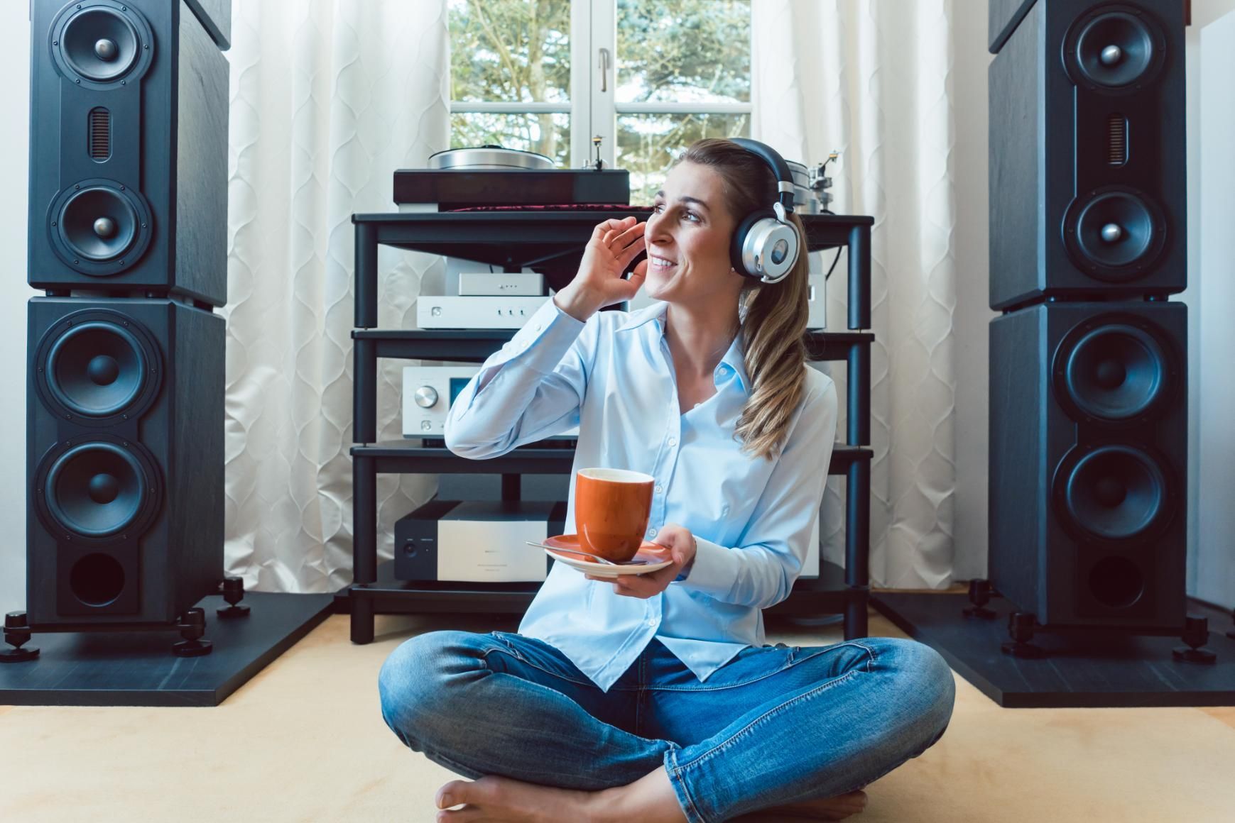 Woman sitting cross-legged with headphones, holding a mug, between large speakers in a bright room
