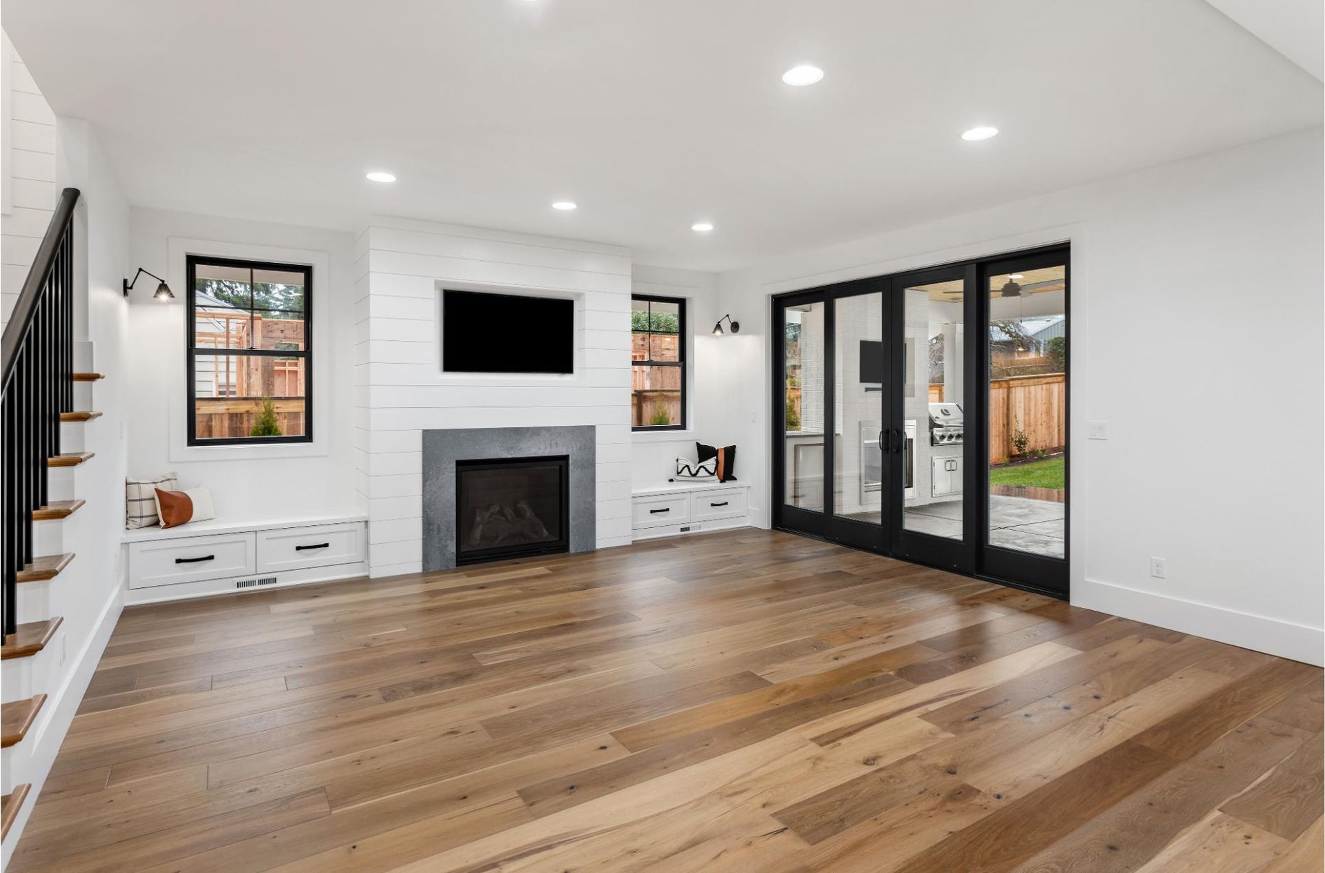Bright living room with hardwood floors, white fireplace, staircase, and black-framed glass doors