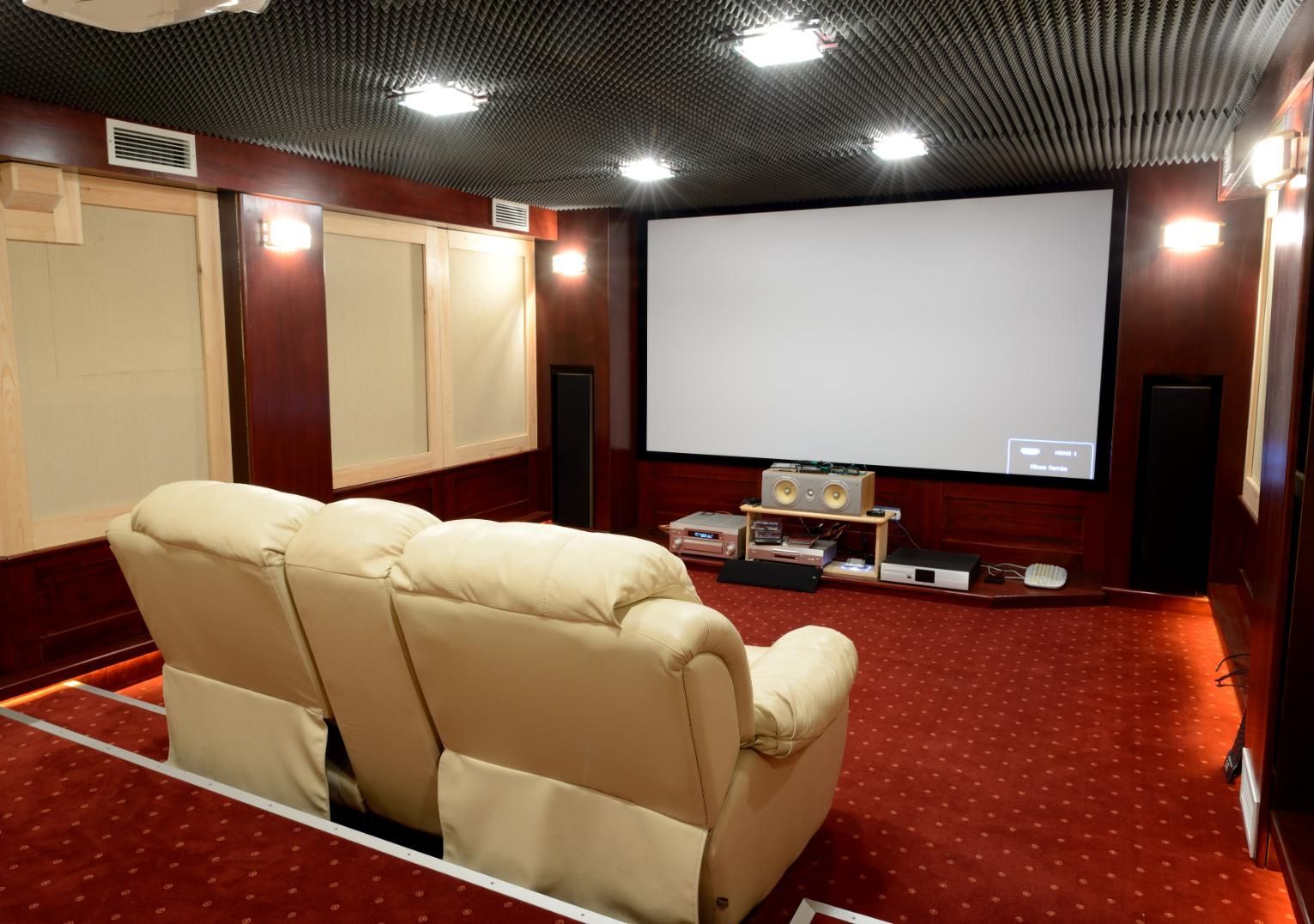 Home theater room with beige recliners facing a large projection screen and red carpet.