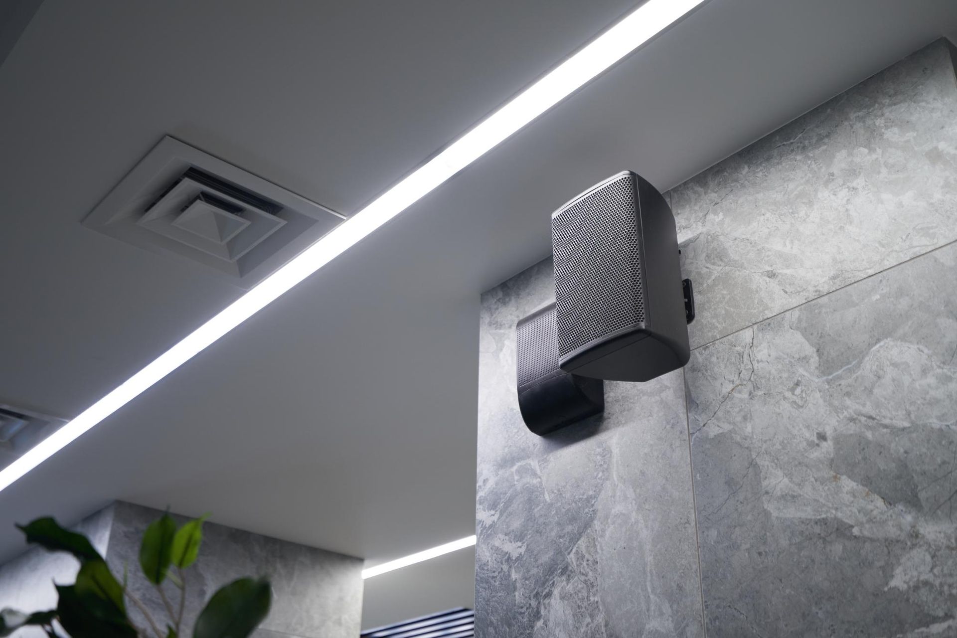 Wall-mounted speaker on a gray tiled wall beneath a linear ceiling light in a modern interior