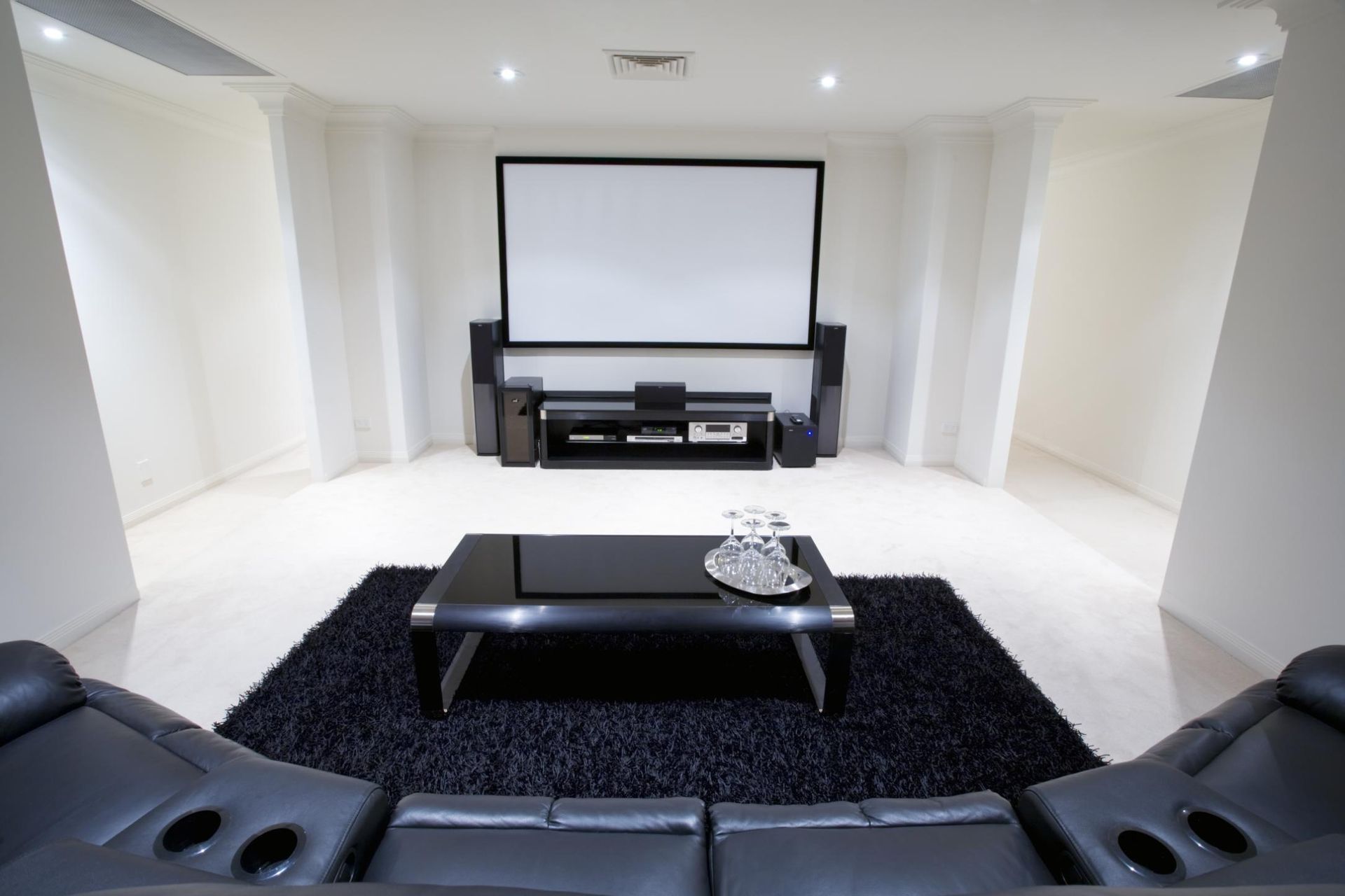 Modern home theater with projector screen, black leather seats, and a black coffee table on a shag rug.