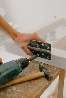 Person using a drill to attach a hinge to a white wooden frame on a wooden work surface.