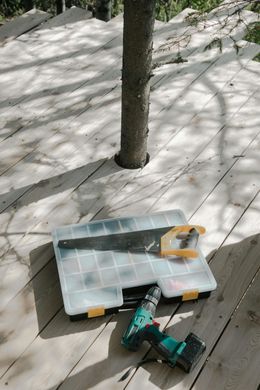 Saw, drill, and tool case on a wooden deck, surrounding a tree trunk. Sunny outdoor setting.