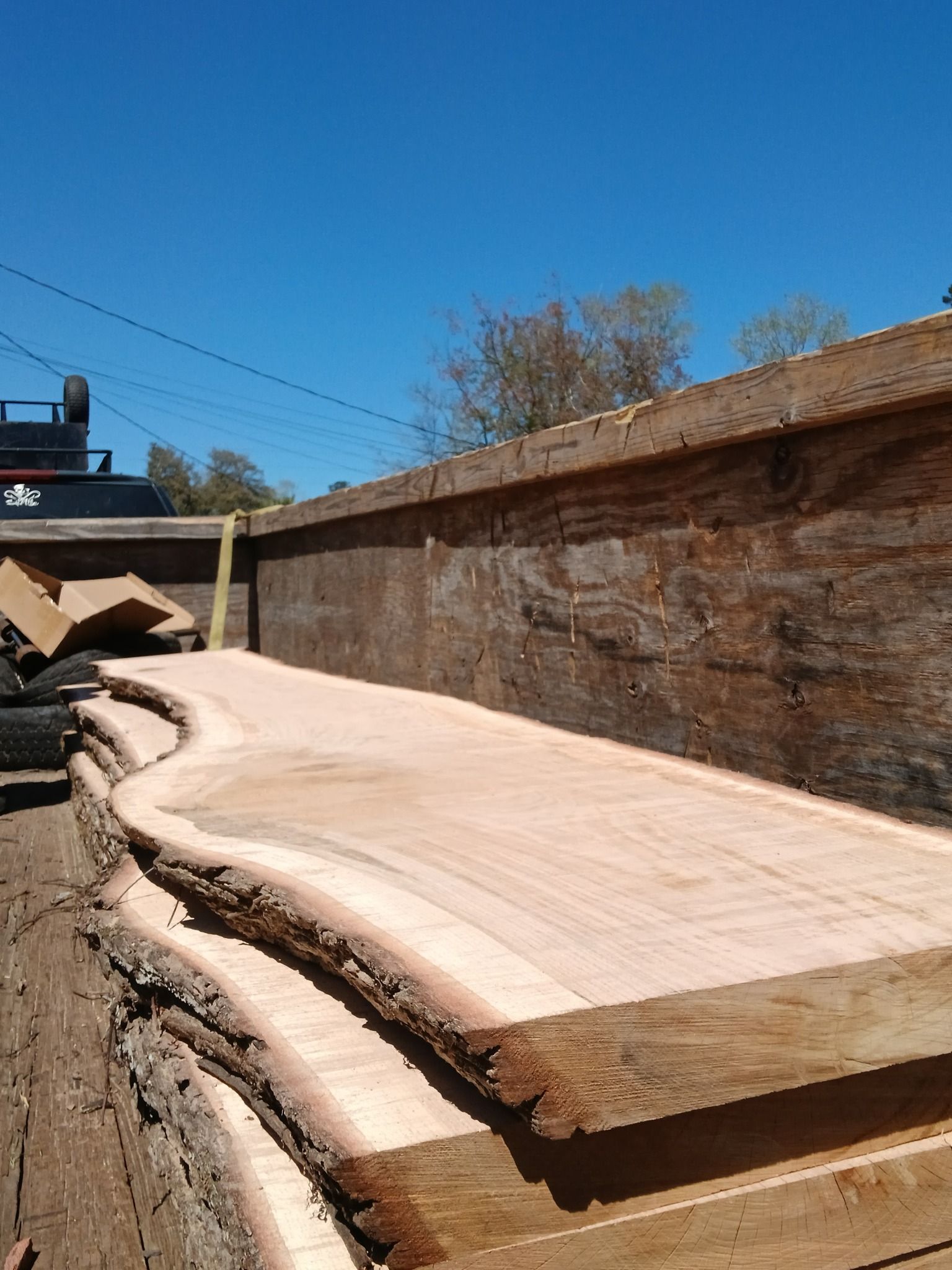 A large, unfinished wooden slab with live edges sits in a wooden container under a blue sky.