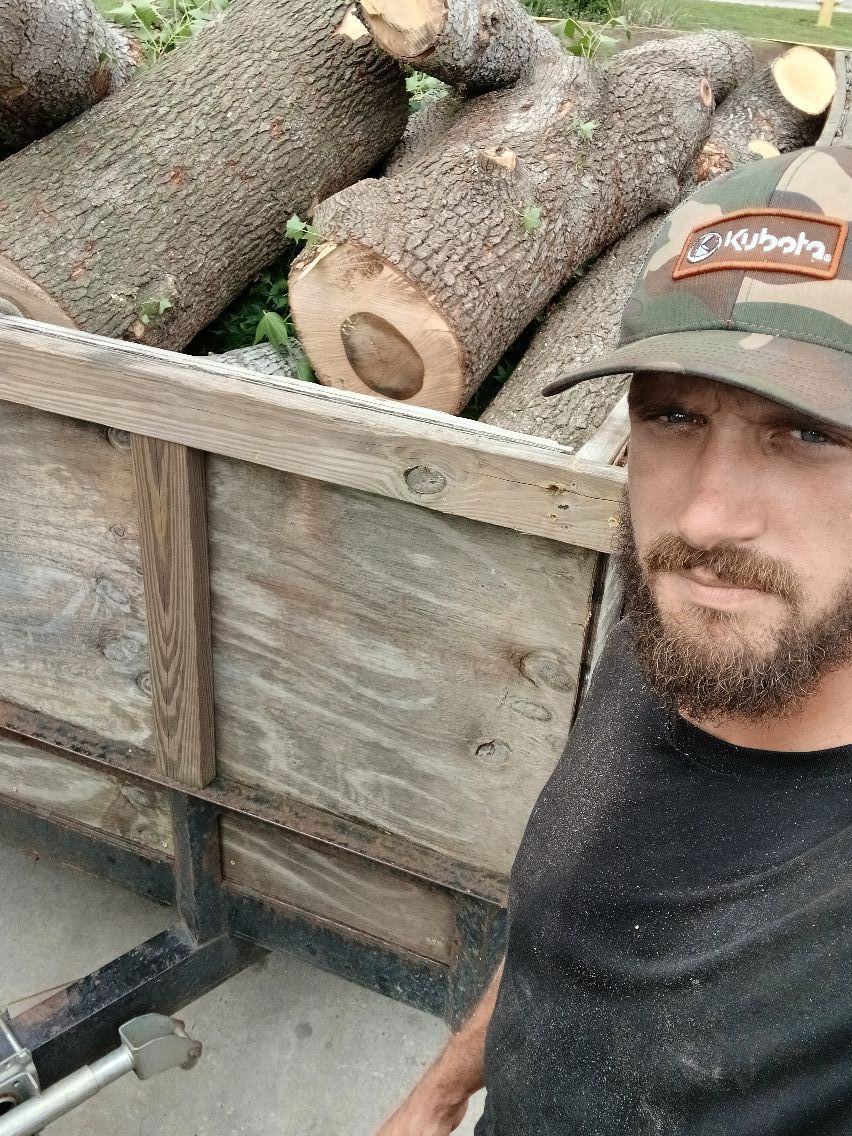 Man with beard and camouflage hat by a trailer loaded with cut logs.