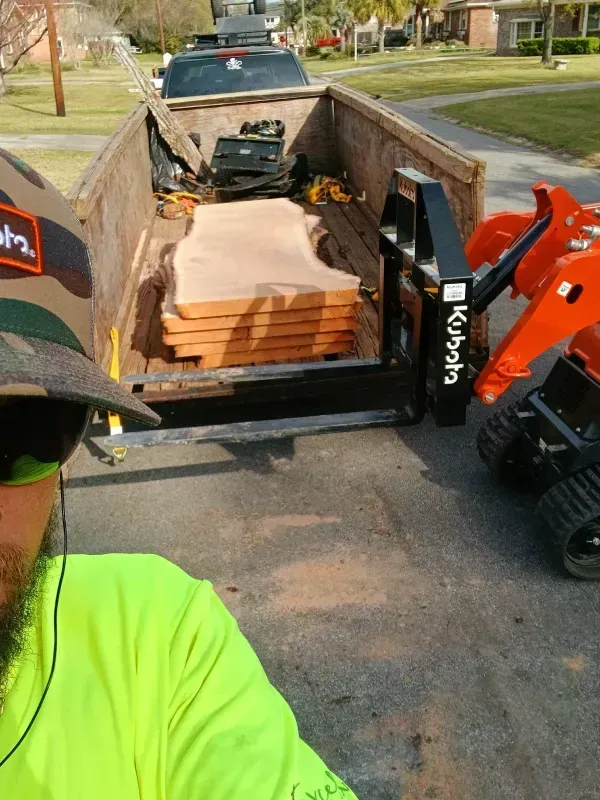 Man in neon shirt near a trailer loaded with wooden slabs, Kubota tractor visible.