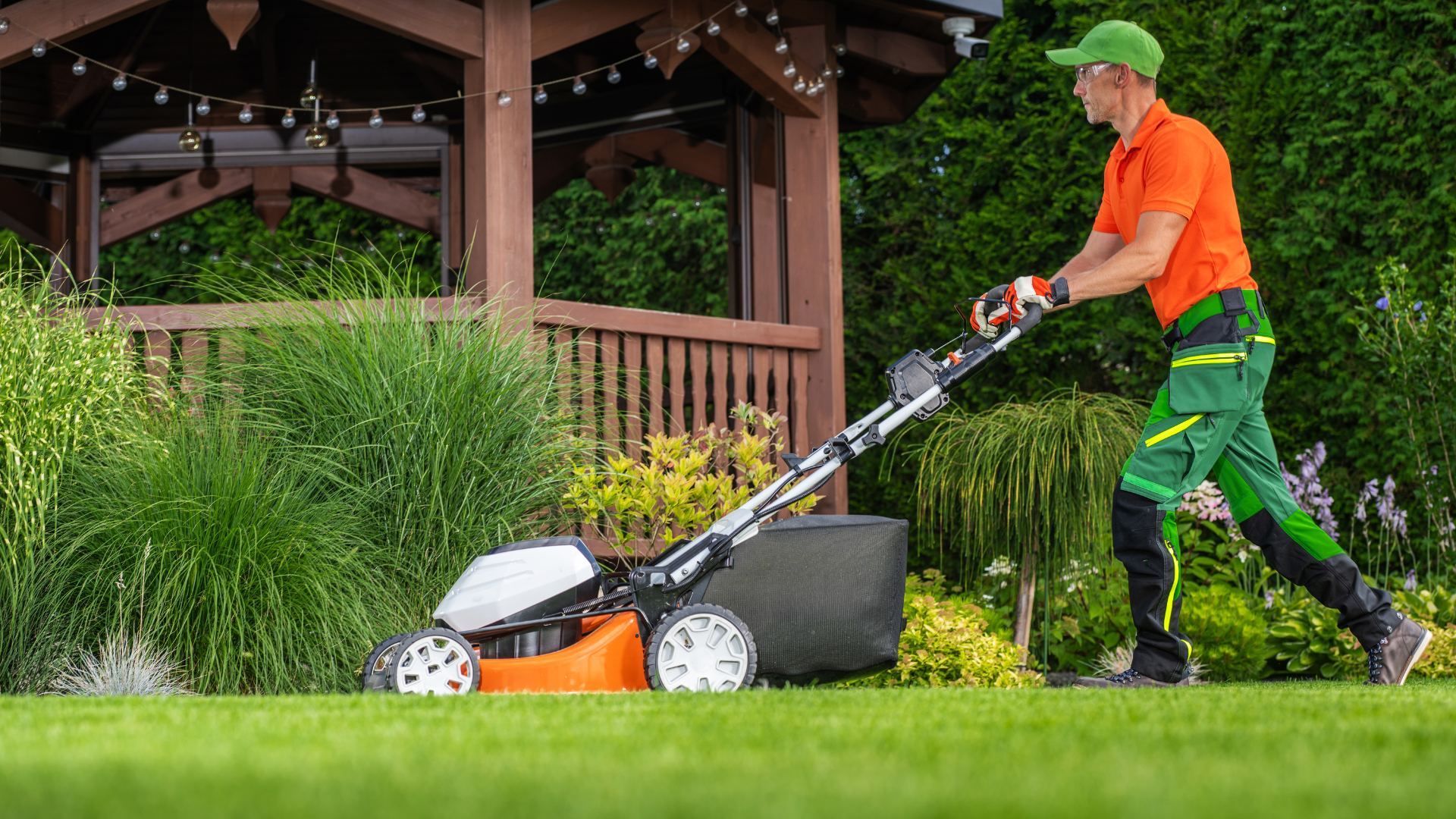 Man mowing lawn with an orange and black mower, wearing orange shirt, green pants and hat in a garden setting.