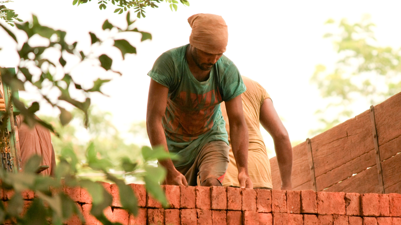Man and person building a brick wall outdoors, wearing work clothes, near trees.