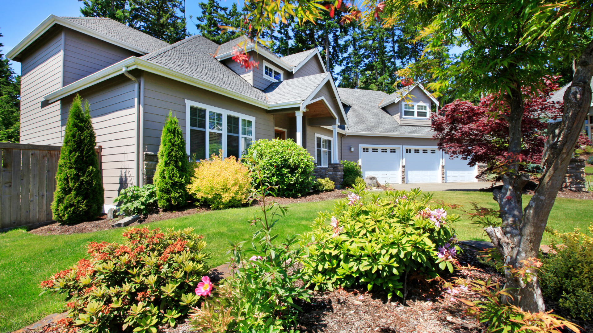 Beige house with green lawn and colorful bushes under a blue sky.
