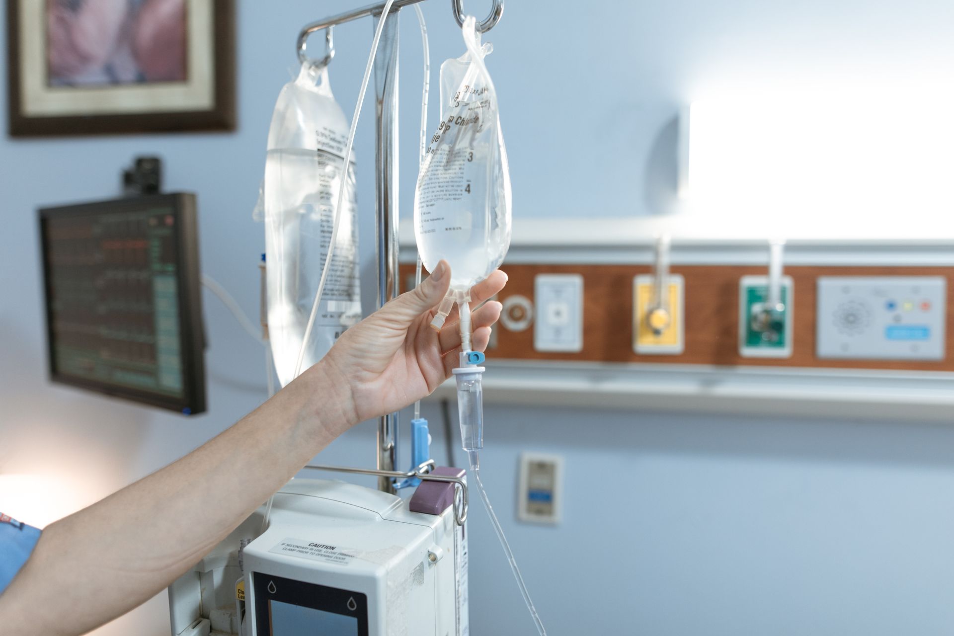 Hand adjusting IV drip bags beside a hospital bed monitor