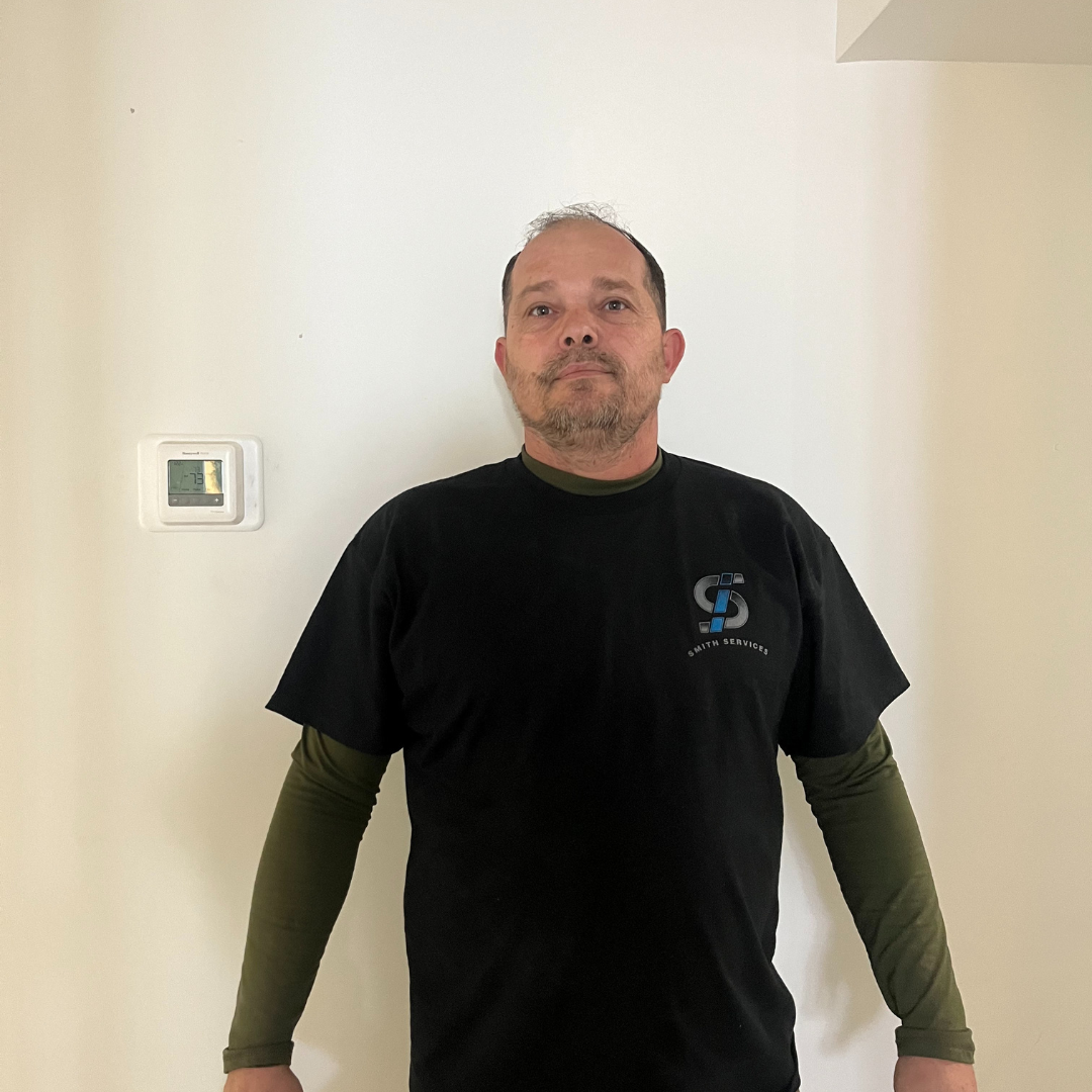 Man in black t-shirt and olive long sleeves stands against a white wall; small white box to his left.