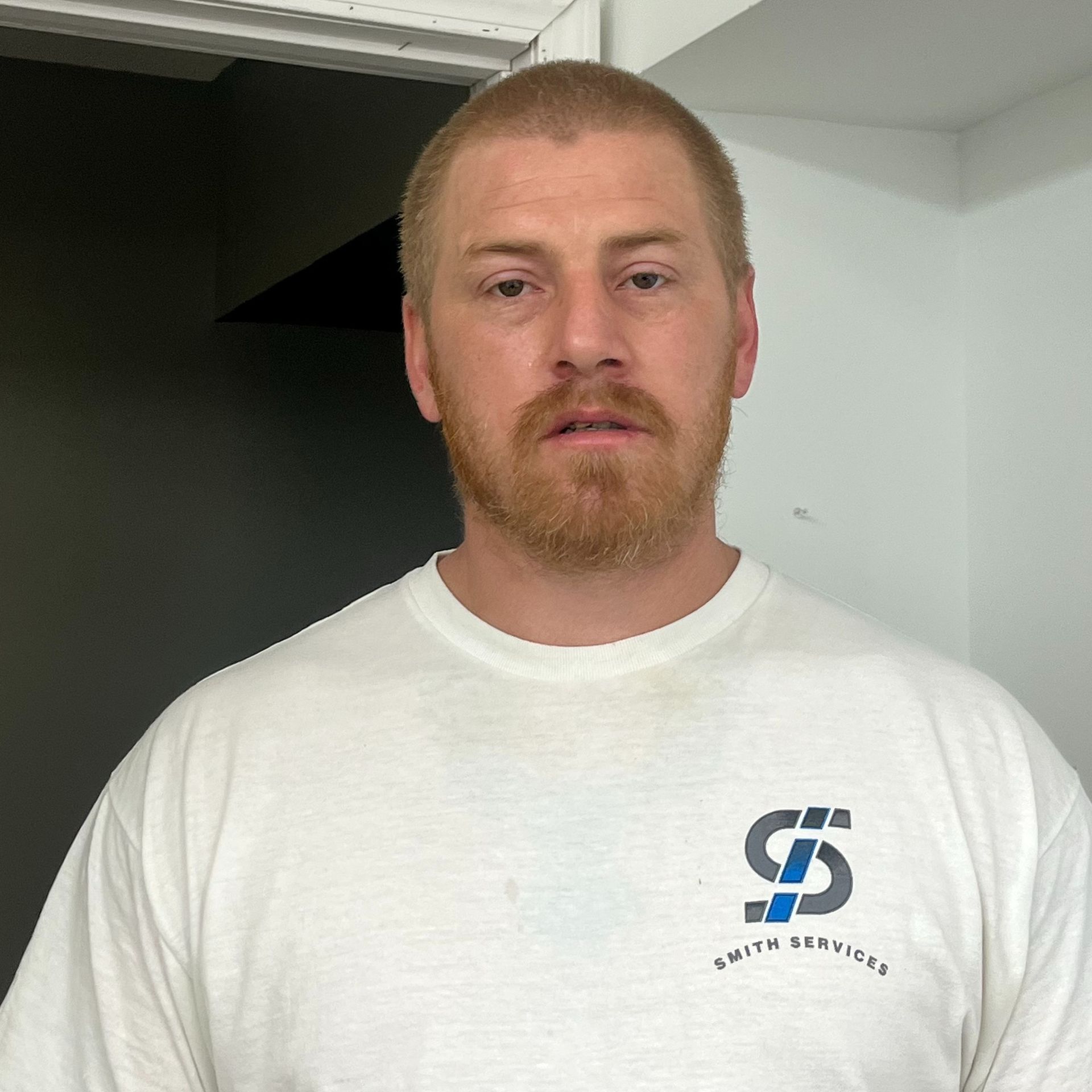 Man in black t-shirt and olive long sleeves stands against a white wall; small white box to his left.