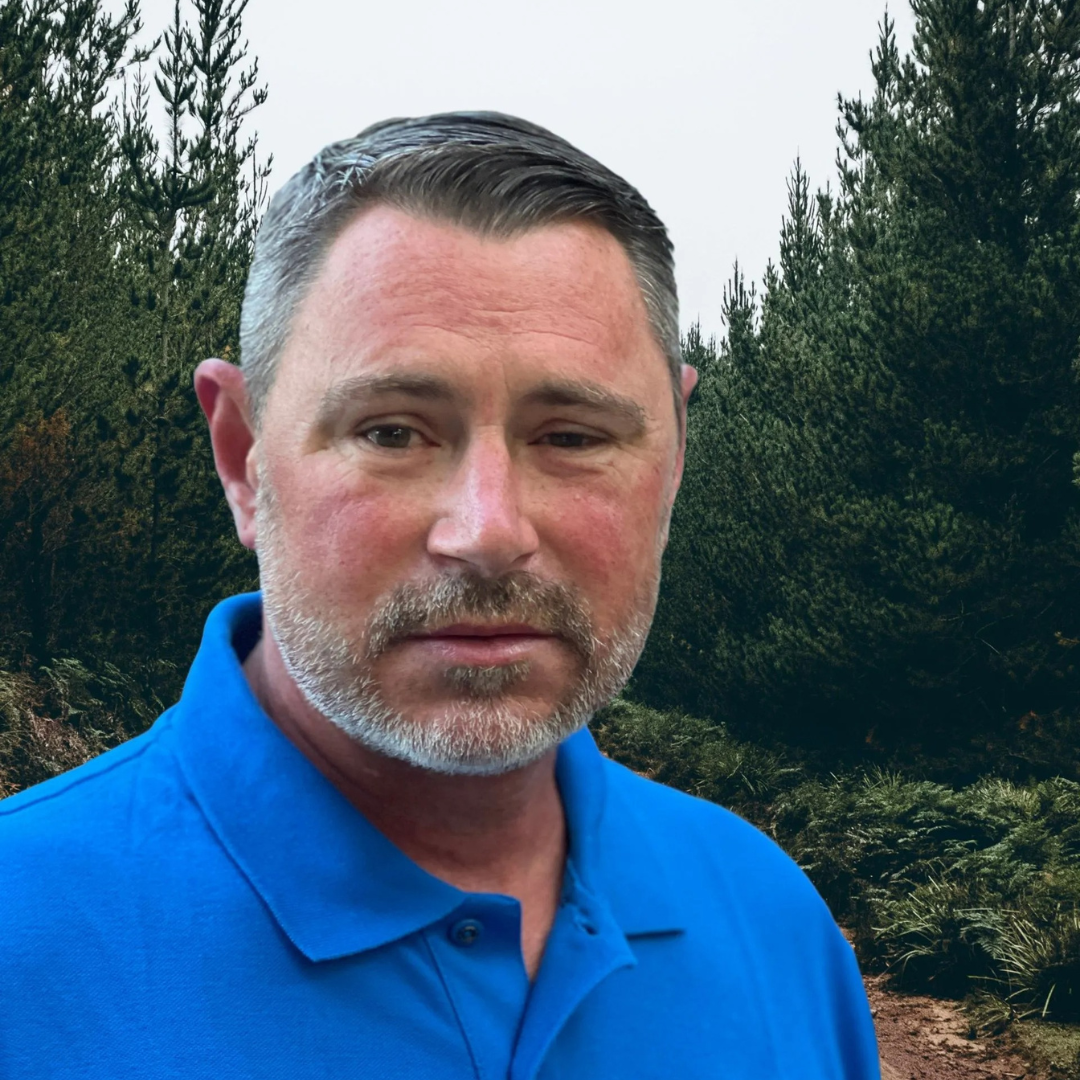 Man in blue shirt, trees in the background, looking at the camera.