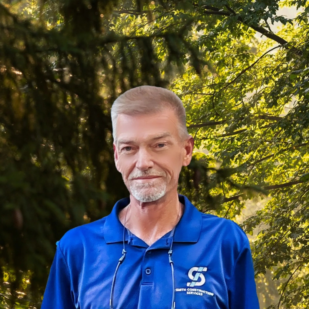 Man in blue shirt, light skin, short gray hair, smiling, in front of a blurred green forest backdrop.