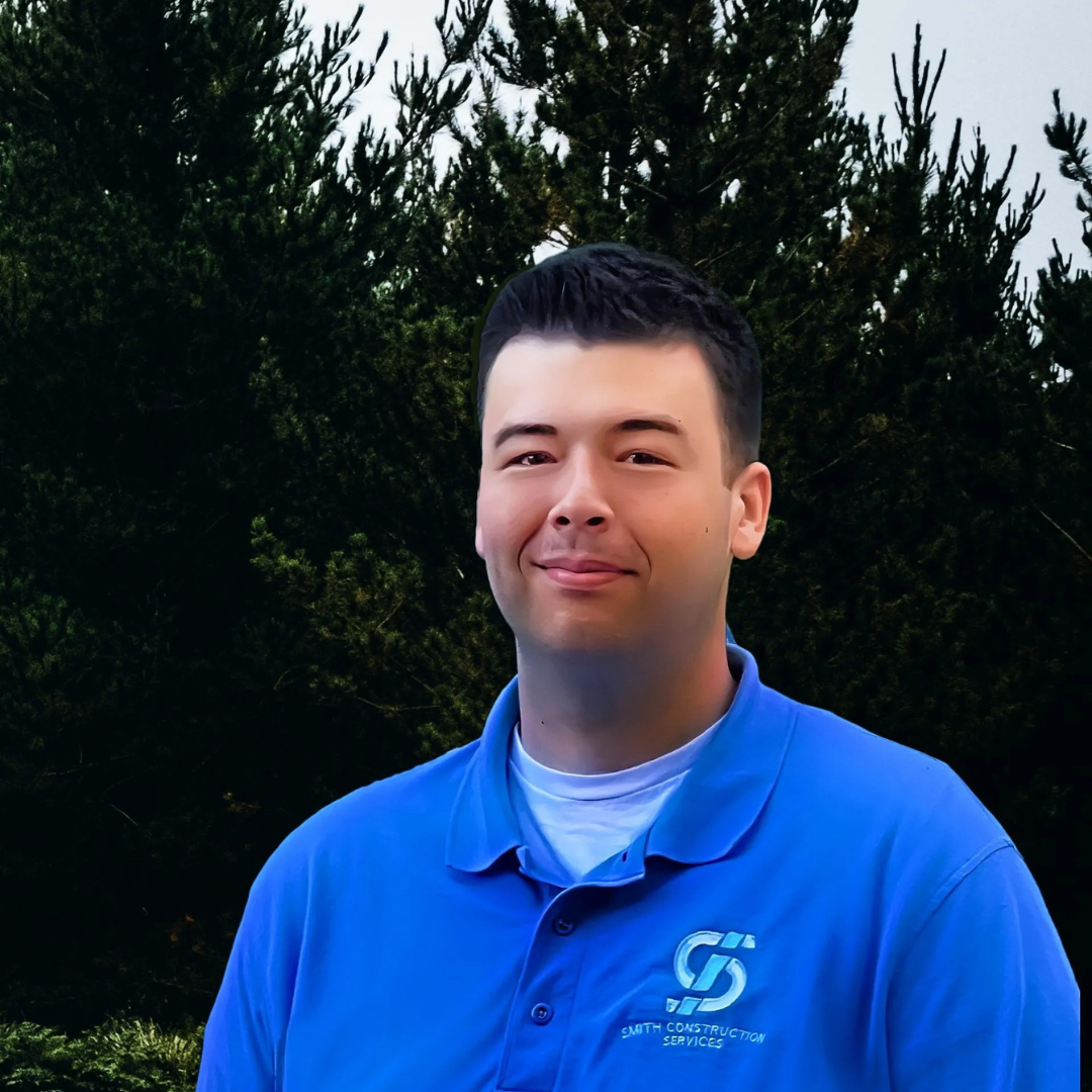 Man in blue polo shirt smiles in front of a green tree.