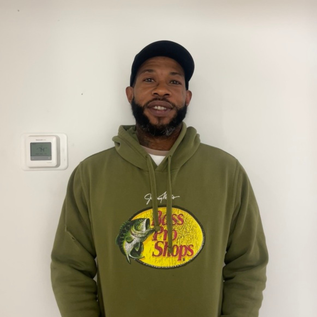 Man wearing a black cap and green Bass Pro Shops hoodie smiles in front of a white wall.
