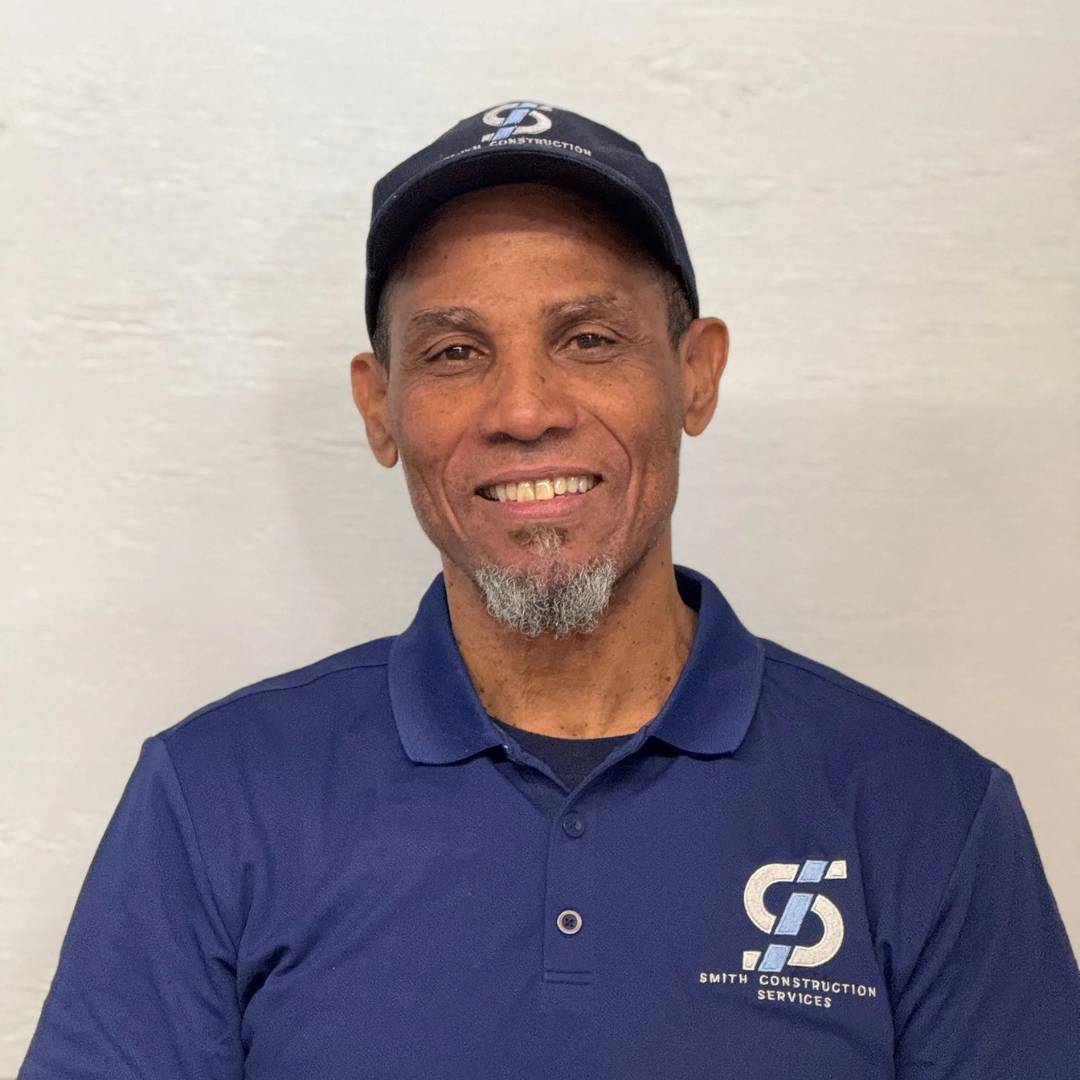 Man wearing a blue cap and polo shirt smiles at the camera against a plain wall.
