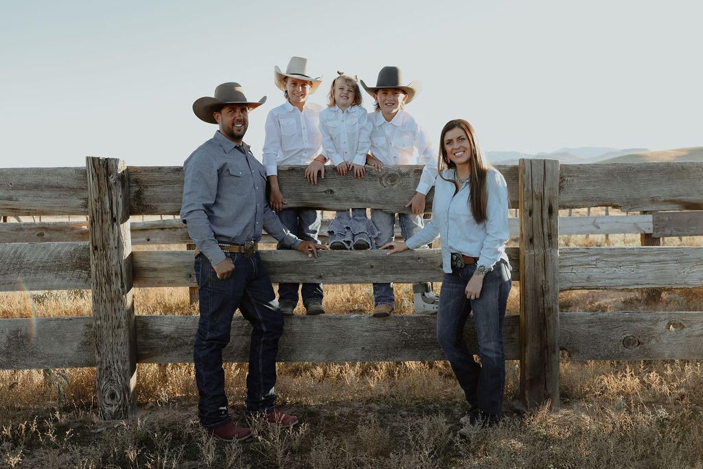 Four people posing by a rustic fence in a dry field, two sitting on the rail and two standing beside them.