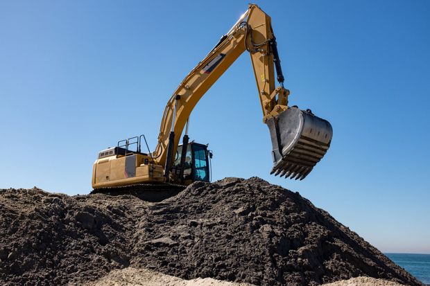 Un escavatore giallo scava un mucchio di terra scura contro un cielo azzurro, vicino all'acqua.