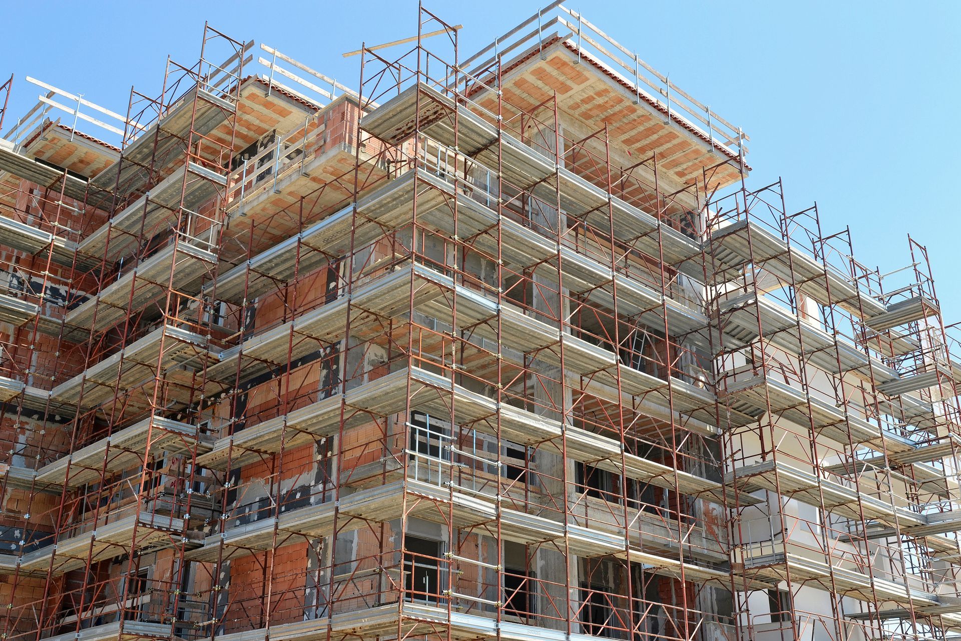 Edificio in costruzione con impalcature, mattoni rosso-marroni e struttura in cemento sotto un cielo azzurro e terso.