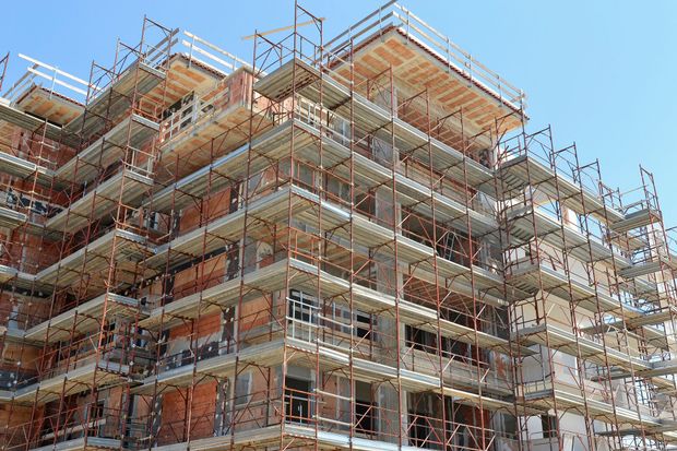 Edificio in costruzione con impalcature, mattoni rosso-marroni e struttura in cemento sotto un cielo azzurro e terso.