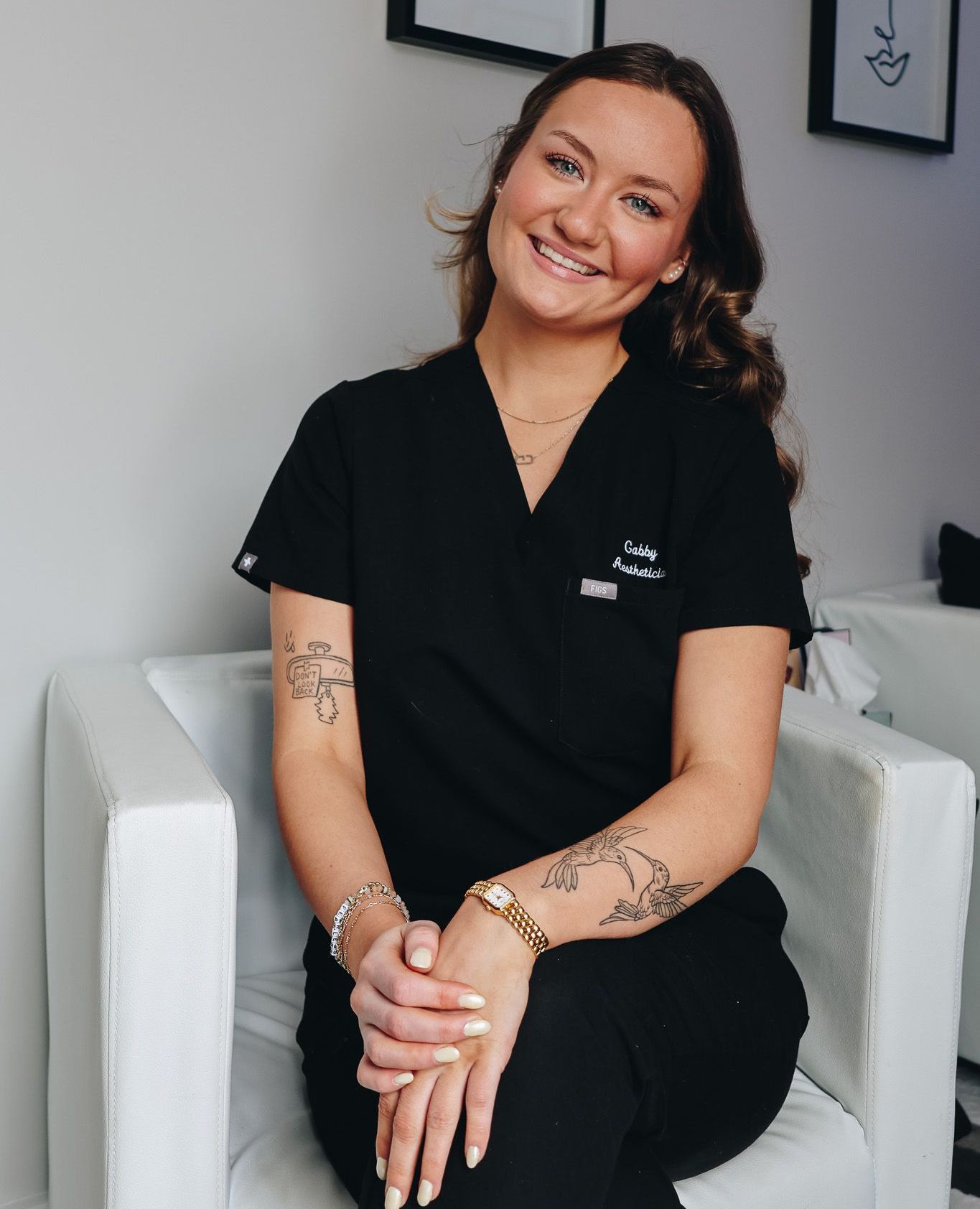 A woman in a black scrub top is sitting in a white chair.