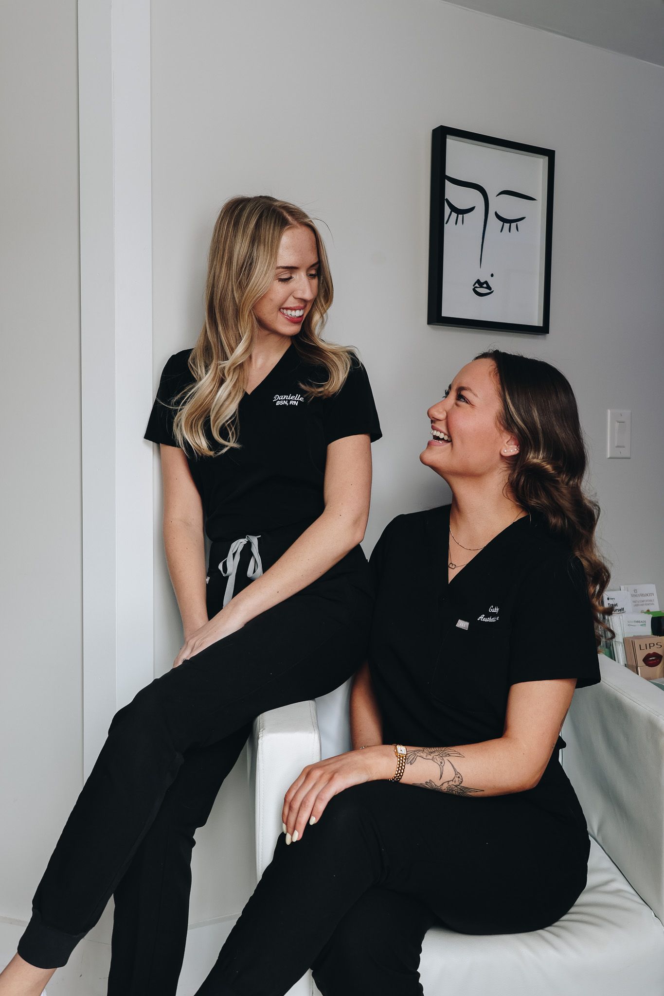 Two women in black scrubs are sitting next to each other.