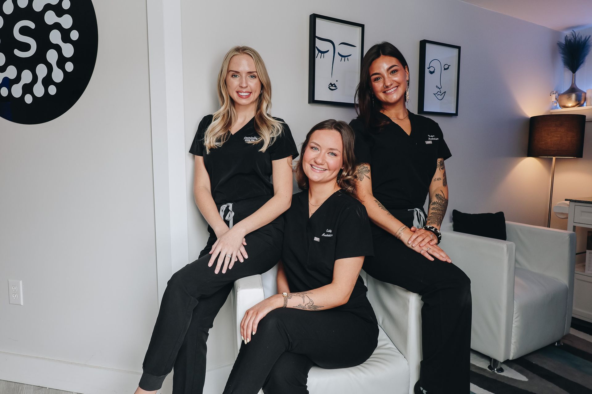 Three women in scrubs are posing for a picture while sitting on a chair.