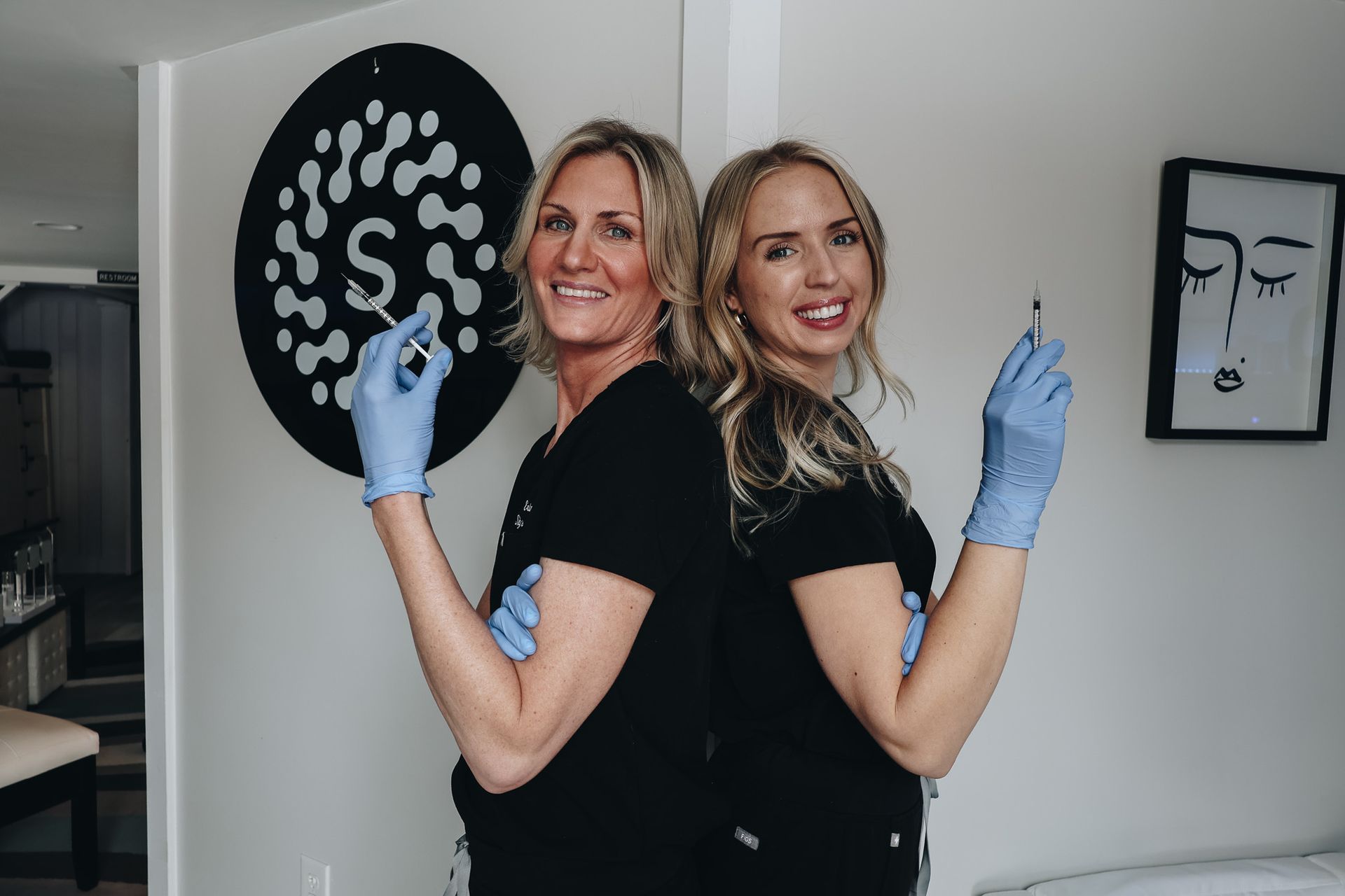 Two women are standing back to back in a room holding syringes.