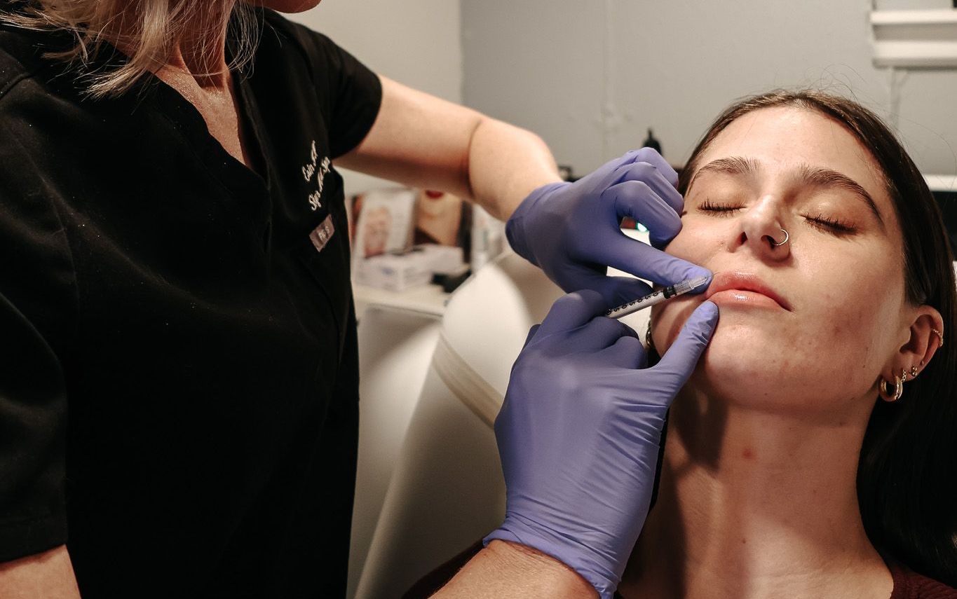 A medical professional in scrubs giving a lip injection to a young woman. The woman is seated in a chair.