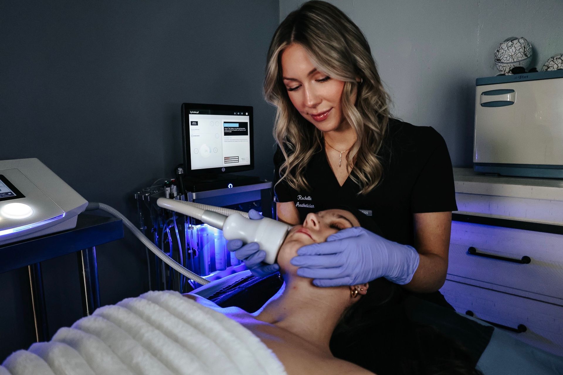 Aesthetian performing a facial treatment on a client. She wears gloves, the client is lying down, and there is medical equipment visible.