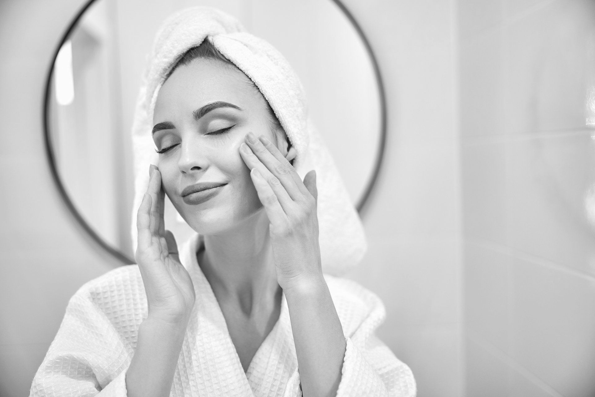 A woman with a towel wrapped around her head is washing her face in front of a mirror.