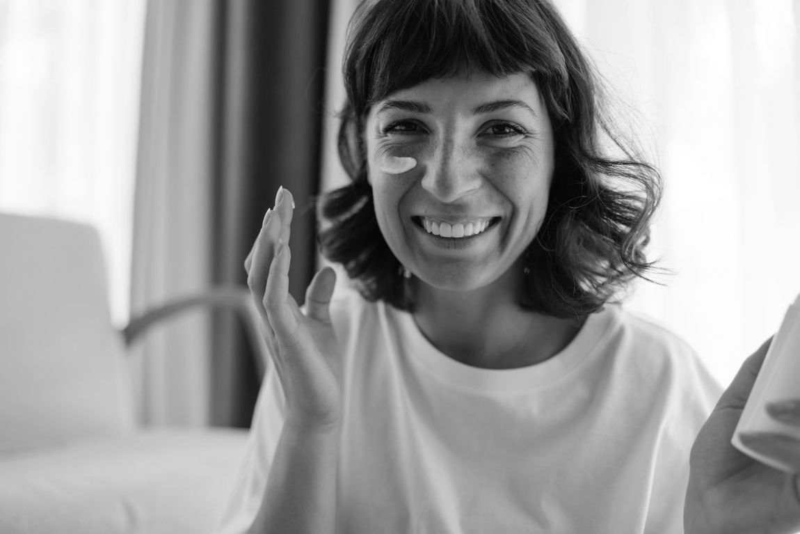 A woman is smiling while applying cream to her face.