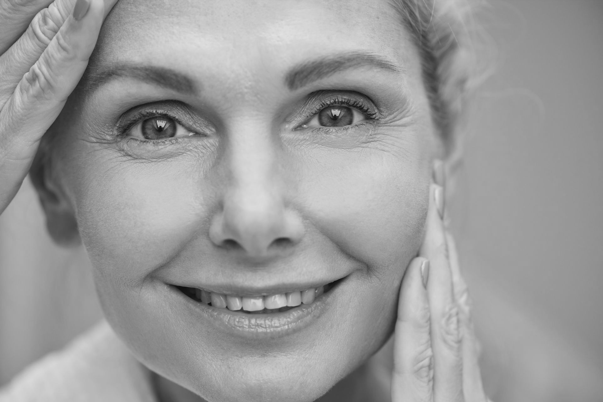 A woman is smiling and touching her face in a black and white photo.