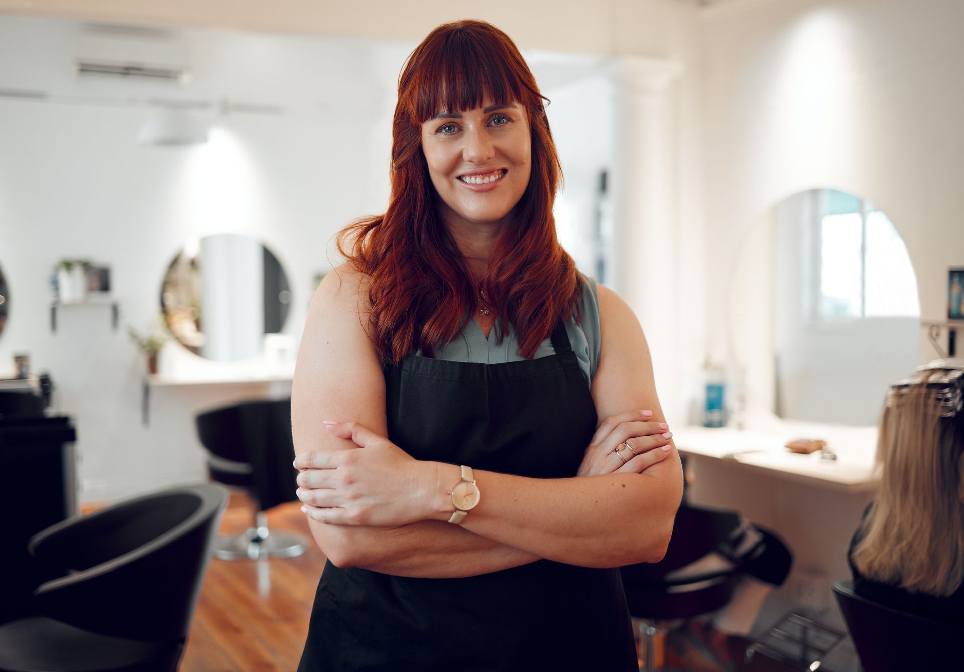 A woman is standing in a salon with her arms crossed and smiling.