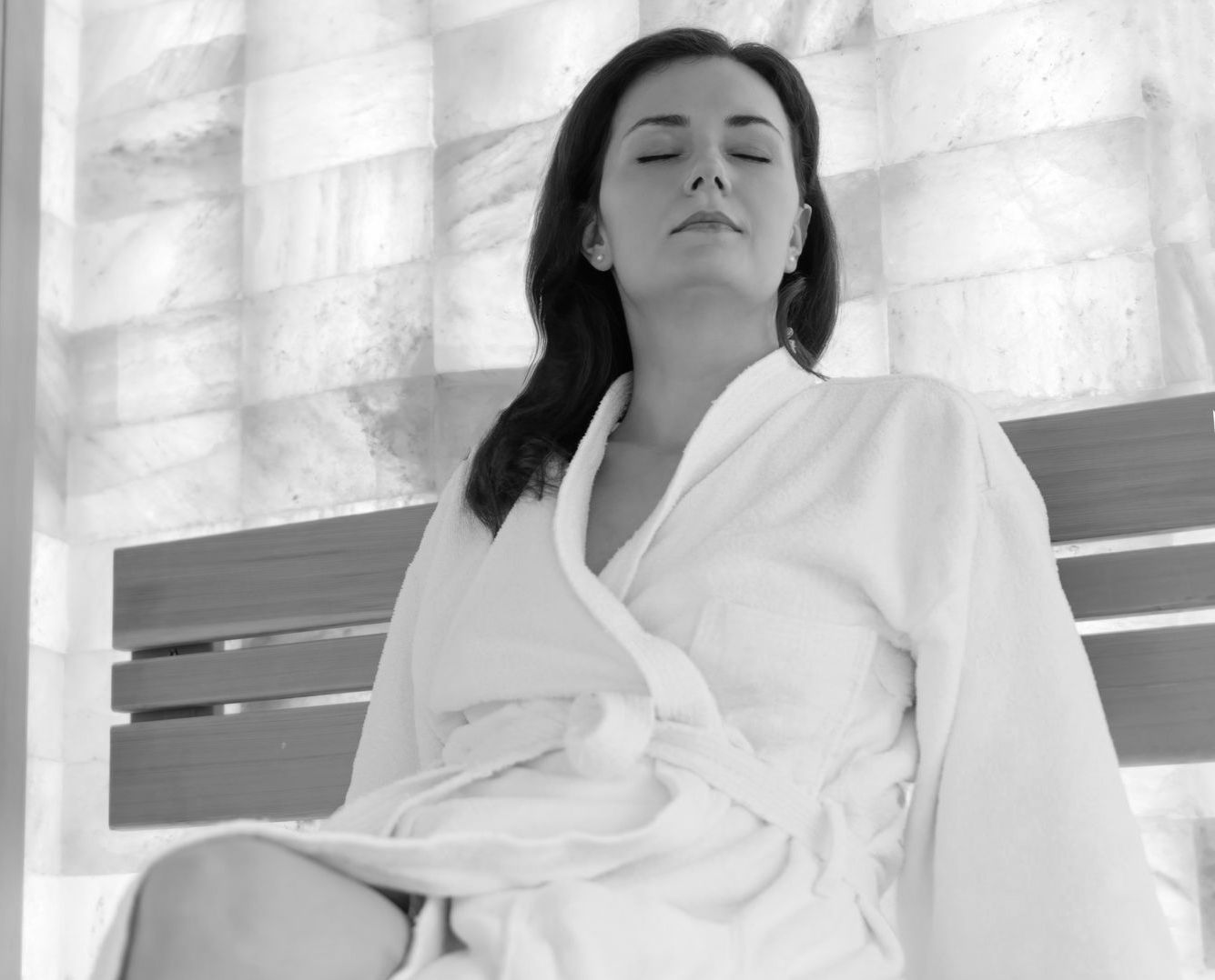 A woman in a bathrobe is sitting on a bench with her eyes closed.