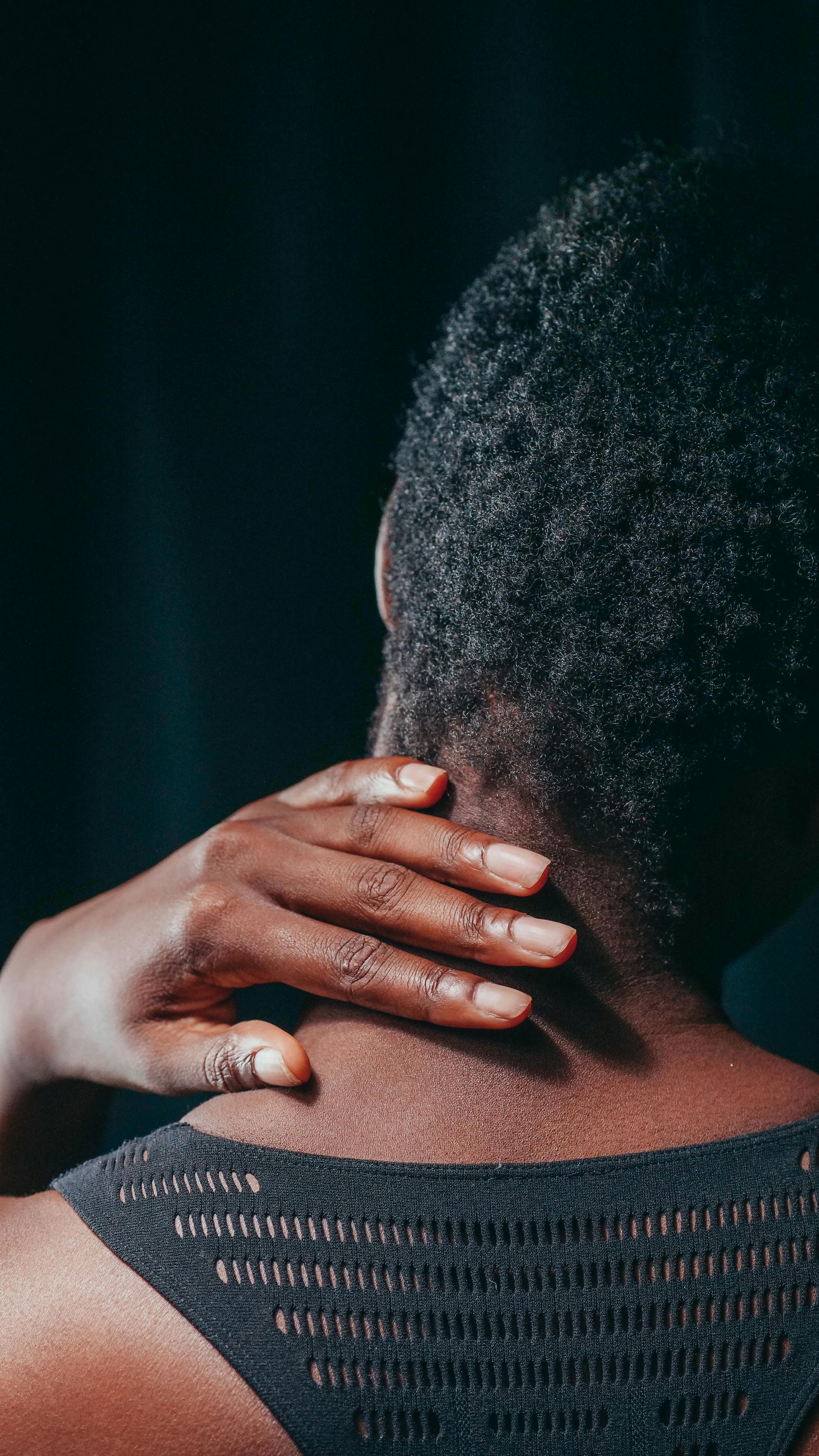 A person touching the back of their neck against a dark background, experiencing neck pain. 