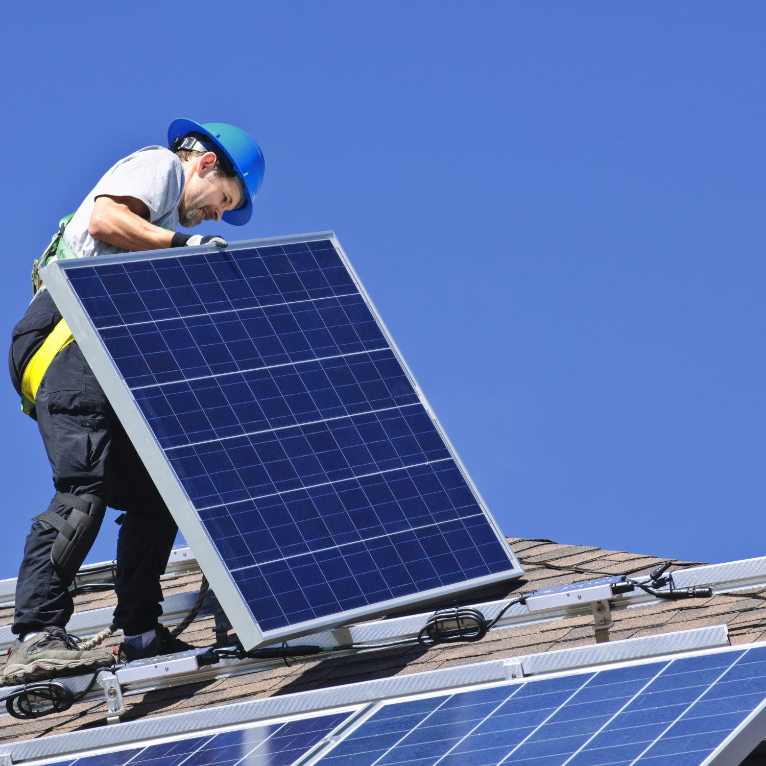 Person in blue hard hat installs solar panel on a rooftop with clear blue sky in the background.