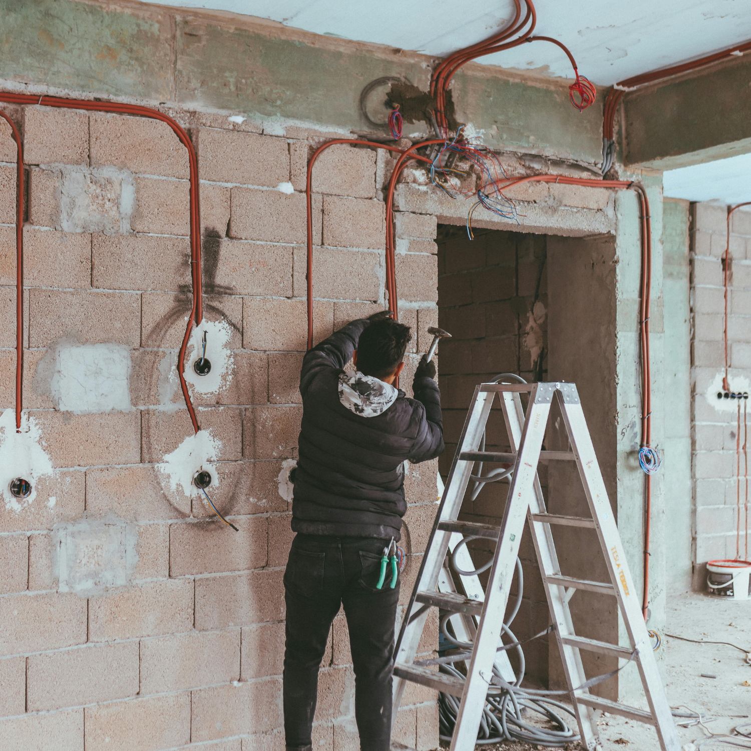 Construction worker installing electrical wiring on a brick wall, using a ladder and tools.