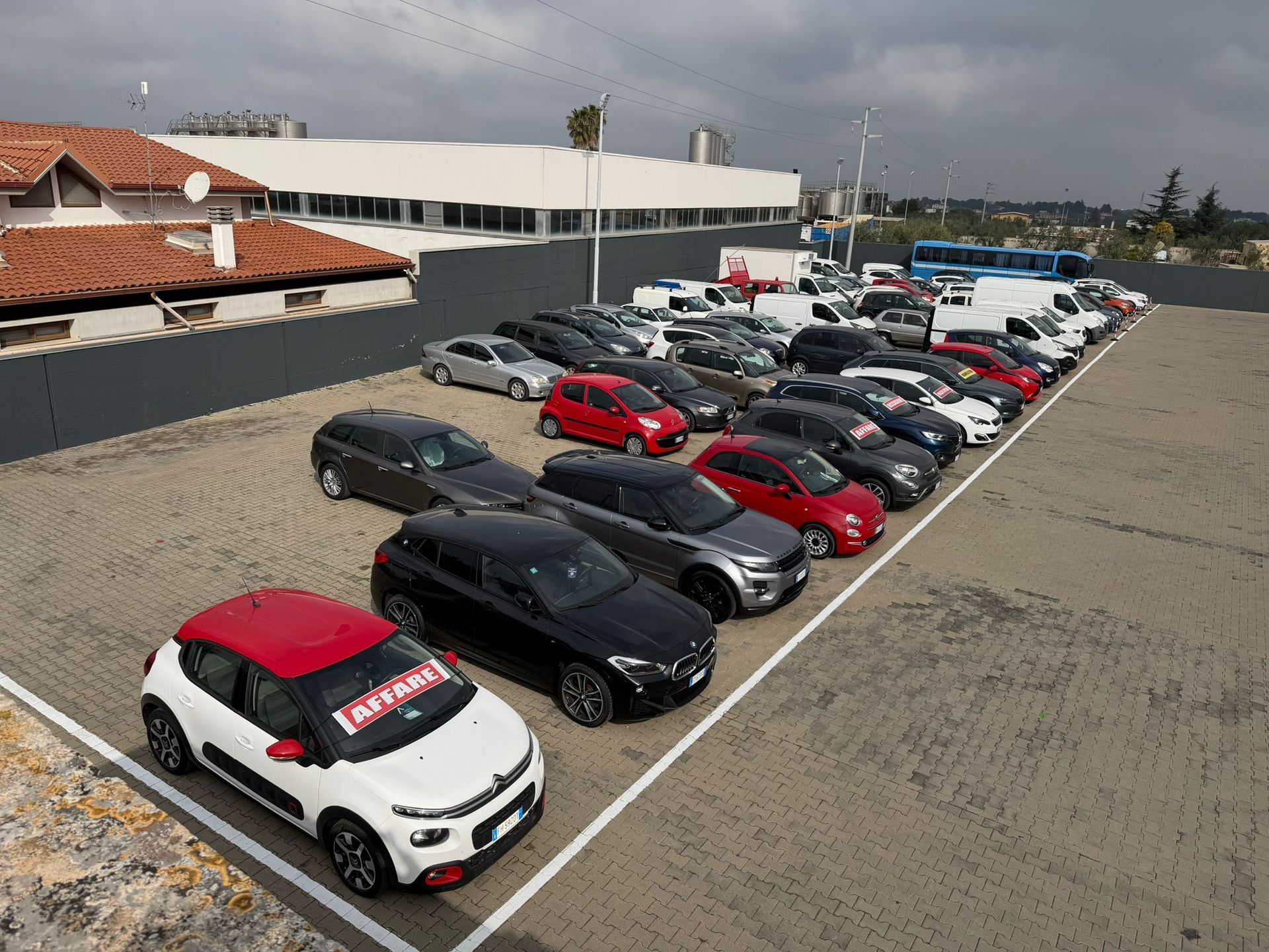File di auto parcheggiate accanto a un edificio bianco in un ampio parcheggio all'aperto sotto un cielo nuvoloso.